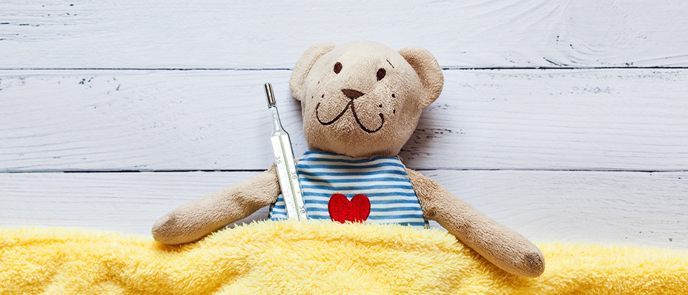 teddy bear tucked into yellow blanket and holding a thermometer